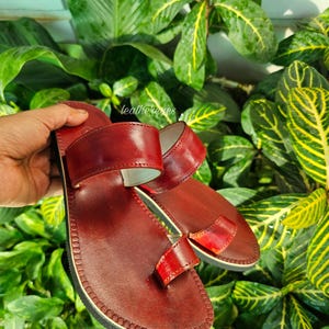 May include: A pair of handcrafted, burgundy leather sandals with a toe loop and double straps. The sandals have a stitched sole and are displayed against a backdrop of lush green foliage.