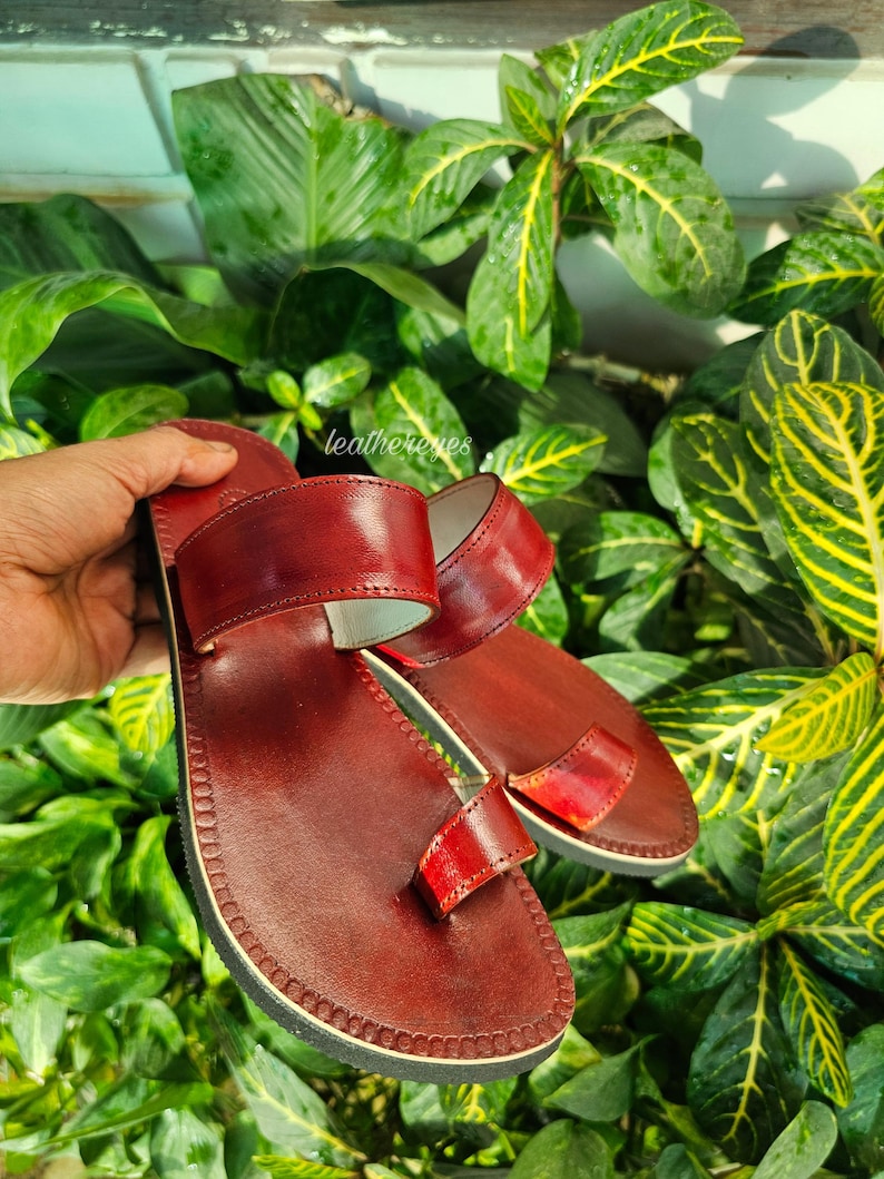 May include: A pair of handcrafted, burgundy leather sandals with a toe loop and double straps. The sandals have a stitched sole and are displayed against a backdrop of lush green foliage.