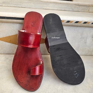 May include: A pair of red leather sandals with a toe loop and a wide strap across the top. The sandals have a dark sole with the text "leathereyes" printed on the sole. The sandals are size 38.