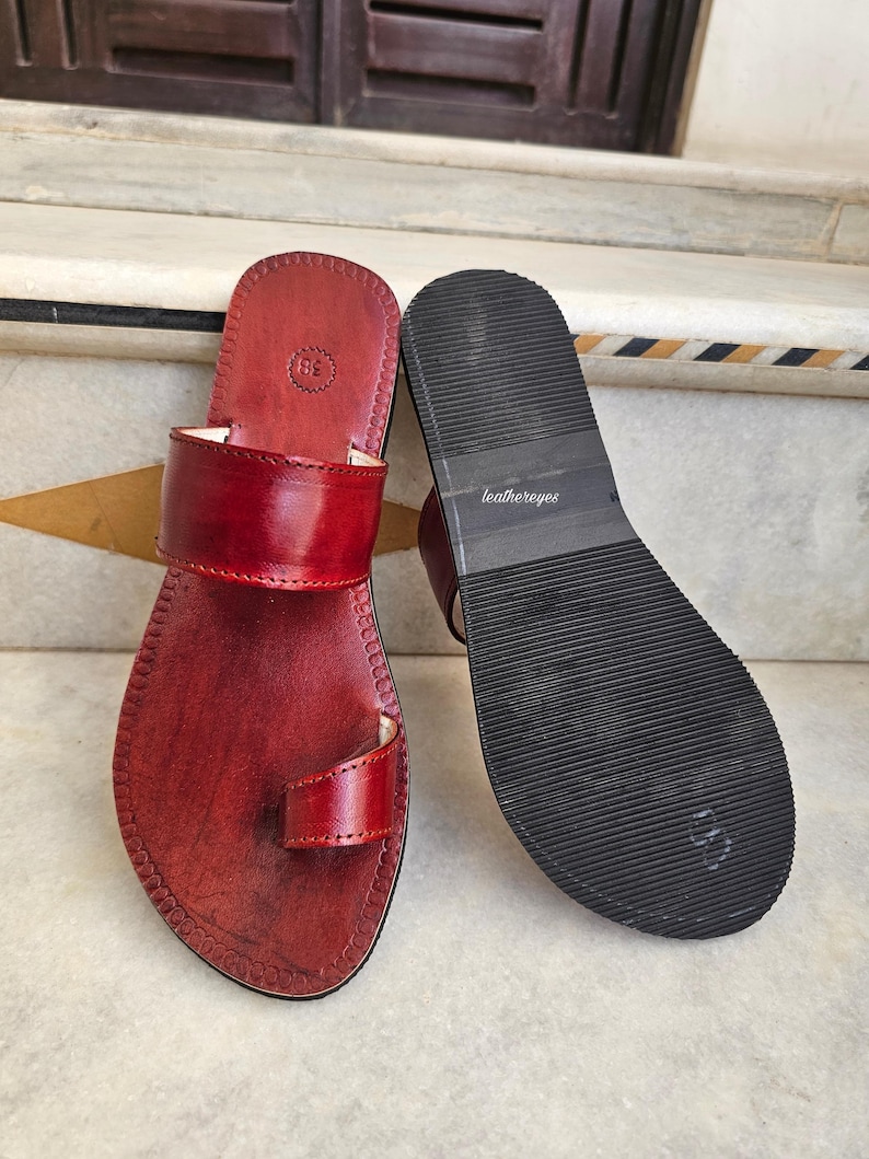 May include: A pair of red leather sandals with a toe loop and a wide strap across the top. The sandals have a dark sole with the text "leathereyes" printed on the sole. The sandals are size 38.