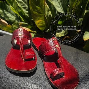May include: A pair of handmade red leather sandals with a toe loop design. The sandals feature decorative stitching and small gold-colored studs. The image includes the text "LEATHEREYES BY RISHI SHOES" and "THIS PRODUCT IS 100% HANDMADE".
