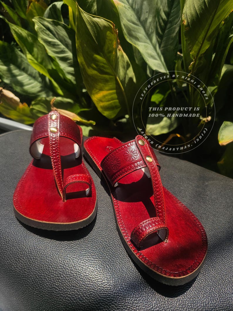 May include: A pair of handmade red leather sandals with a toe loop design. The sandals feature decorative stitching and small gold-colored studs. The image includes the text "LEATHEREYES BY RISHI SHOES" and "THIS PRODUCT IS 100% HANDMADE".