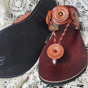 May include: Handmade leather sandals in shades of brown and orange. The sandals feature a decorative floral design on the straps and toe post. The sole is black with a textured pattern. The sandals are displayed on a white lace surface.