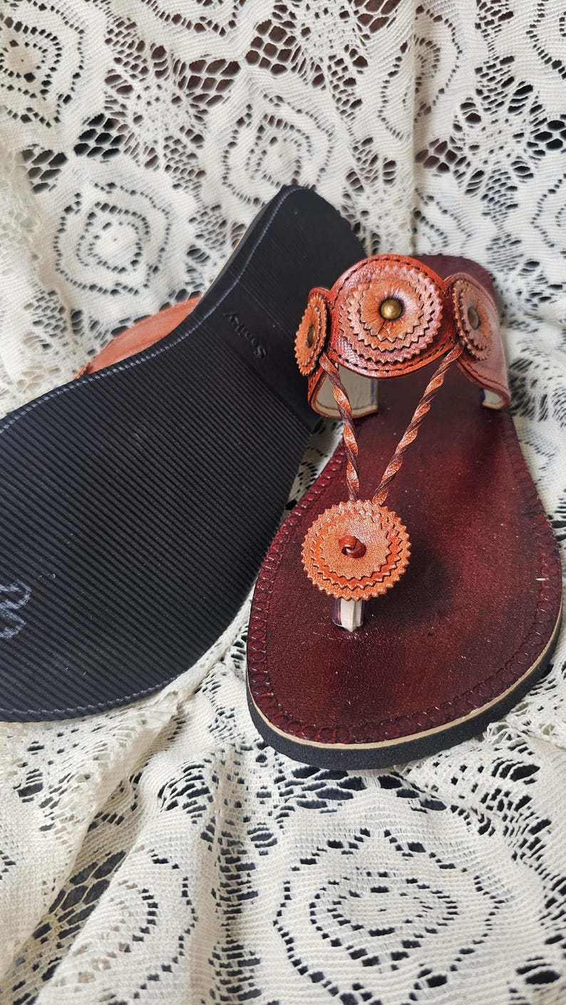 May include: Handmade leather sandals in shades of brown and orange. The sandals feature a decorative floral design on the straps and toe post. The sole is black with a textured pattern. The sandals are displayed on a white lace surface.