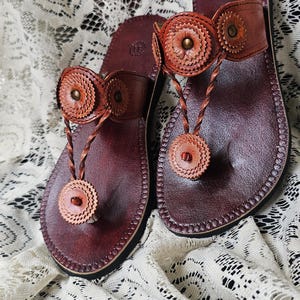 May include: A pair of handcrafted leather sandals. The sandals are a deep burgundy color with decorative, circular accents in a lighter brown. The thong-style straps feature braided details and are designed for comfort and style.