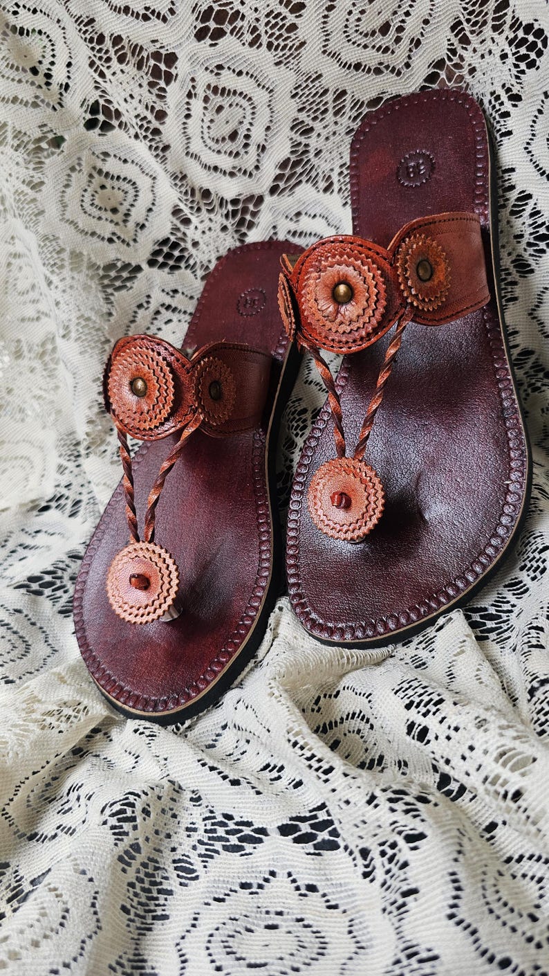 May include: A pair of handcrafted leather sandals. The sandals are a deep burgundy color with decorative, circular accents in a lighter brown. The thong-style straps feature braided details and are designed for comfort and style.