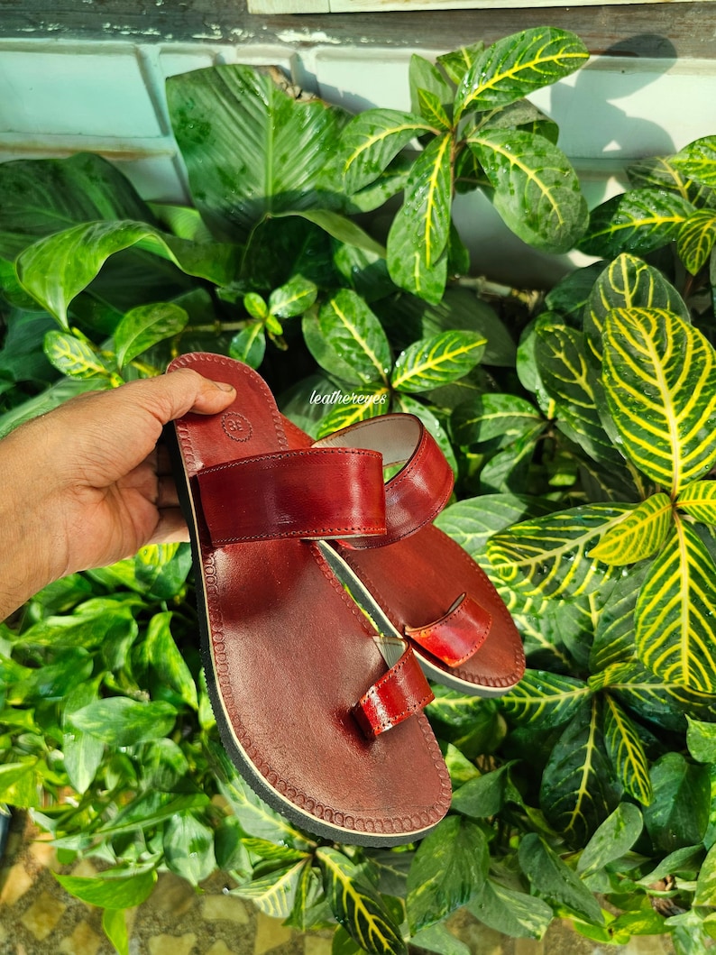May include: A pair of burgundy leather sandals with a toe loop and two straps. The sandals have a stitched sole and are displayed against a backdrop of lush green foliage. The sandals are a size 38.