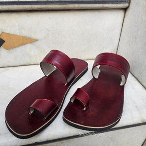 May include: A pair of burgundy leather sandals with a toe loop and a wide strap. The sandals have a dark sole and are displayed on a marble surface. The sandals are a size 9.