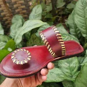 May include: Burgundy leather sandal with gold-coloured accents. The sandal features a circular toe post and a strap across the top of the foot. The design includes decorative stitching and is held by a hand. The brand name "leathereyes" is visible.
