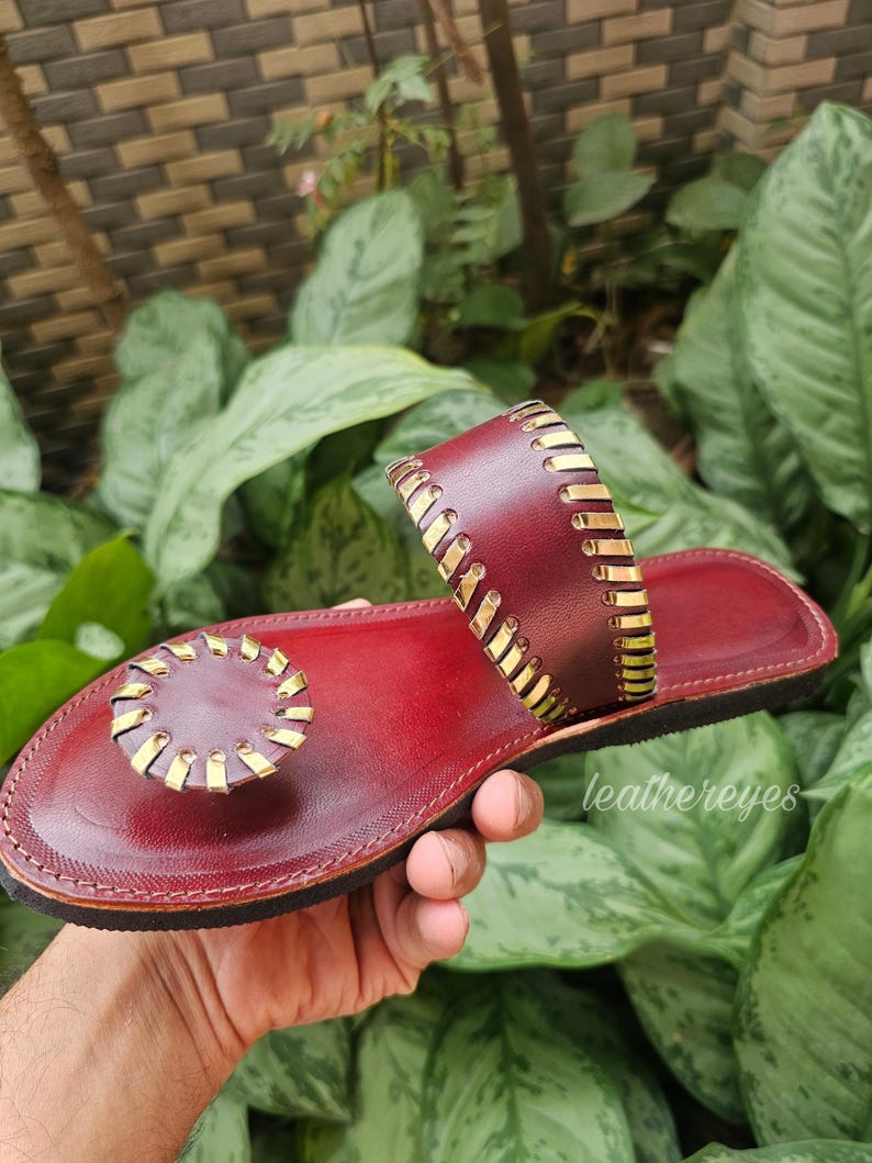 May include: Burgundy leather sandal with gold-coloured accents. The sandal features a circular toe post and a strap across the top of the foot. The design includes decorative stitching and is held by a hand. The brand name "leathereyes" is visible.
