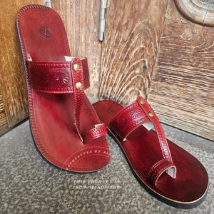 May include: A pair of handmade, red leather sandals. The sandals feature a thong-style design with a strap across the top of the foot, adorned with floral embossing and secured with brass-colored rivets. The soles are black.