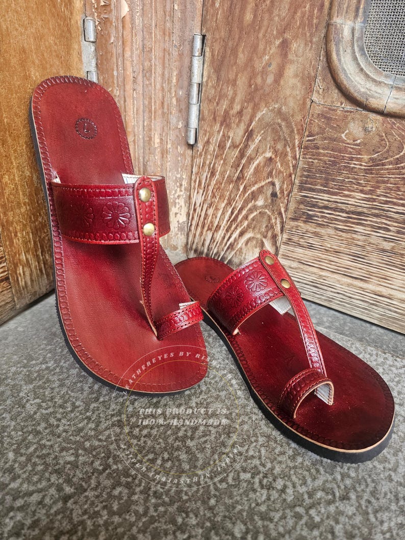 May include: A pair of handmade, red leather sandals. The sandals feature a thong-style design with a strap across the top of the foot, adorned with floral embossing and secured with brass-colored rivets. The soles are black.