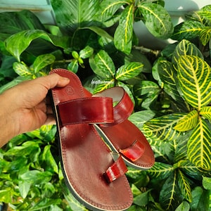 May include: A pair of burgundy leather sandals with a toe loop and two straps. The sandals have a stitched sole and are displayed against a backdrop of lush green foliage. The sandals are a size 38.