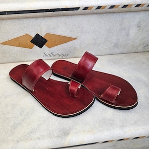 May include: A pair of burgundy leather sandals with a toe loop and a strap across the top. The sandals have a dark sole and a light-colored interior. The sandals are displayed on a marble surface with decorative geometric patterns.