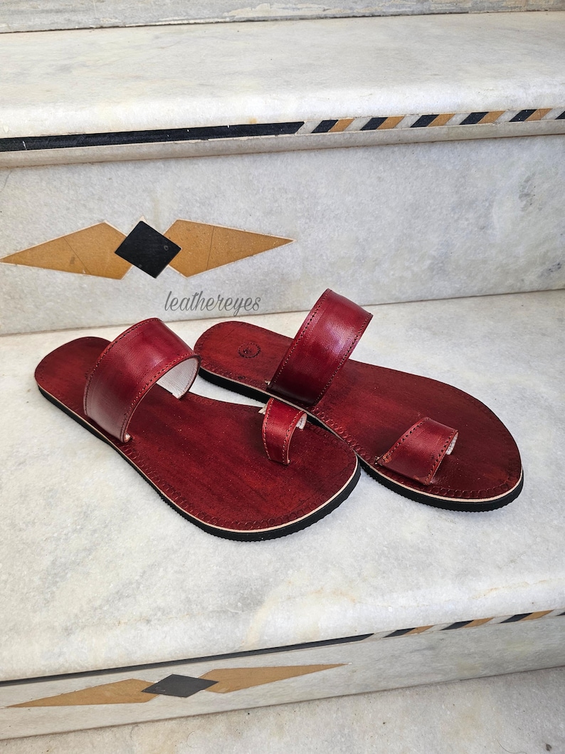 May include: A pair of burgundy leather sandals with a toe loop and a strap across the top. The sandals have a dark sole and a light-colored interior. The sandals are displayed on a marble surface with decorative geometric patterns.