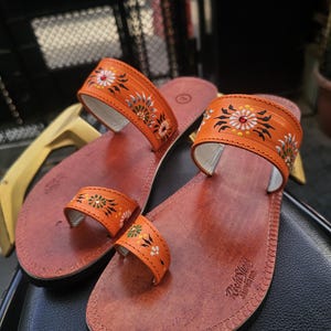Op de afbeelding: Een paar oranje leren sandalen met bloemmotieven. De sandalen hebben een teenband en een band over de bovenkant van de voet, beide versierd met kleurrijke bloempatronen. Het voetbed is roodbruin.