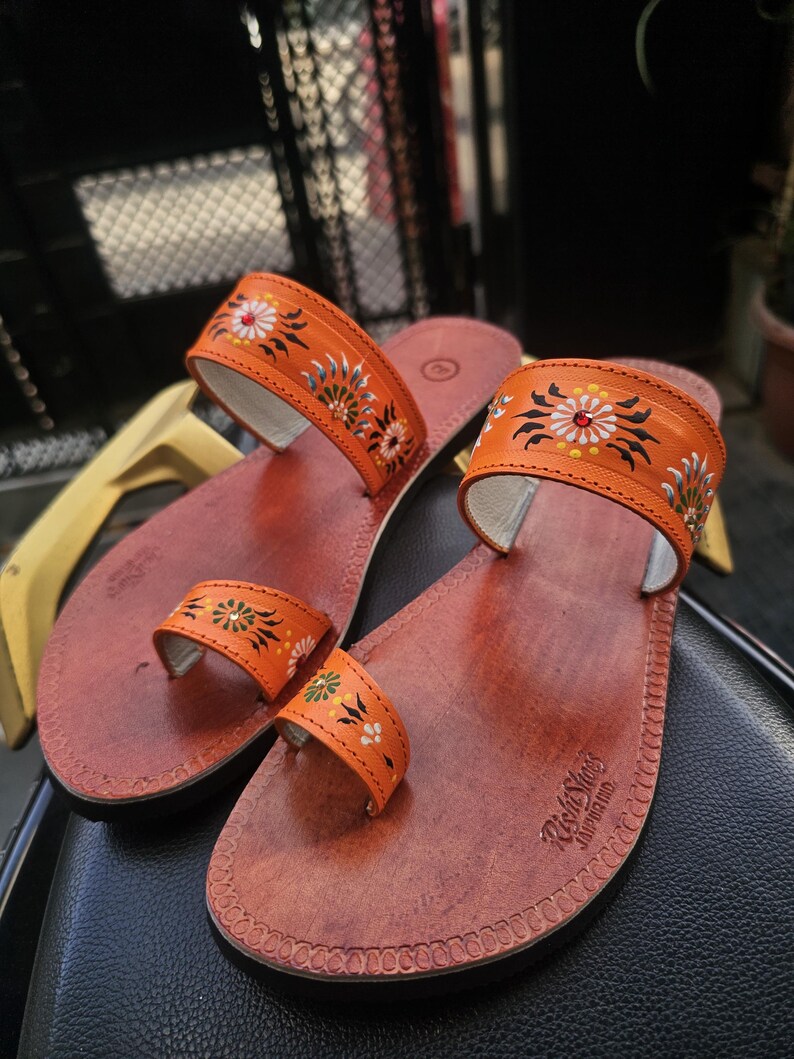 Op de afbeelding: Een paar oranje leren sandalen met bloemmotieven. De sandalen hebben een teenband en een band over de bovenkant van de voet, beide versierd met kleurrijke bloempatronen. Het voetbed is roodbruin.