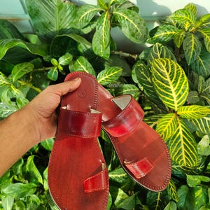 May include: Red leather sandals with a toe loop and a wide strap across the top. The sandals have a stitched edge and a natural leather look. The sandals are displayed against a backdrop of green foliage.