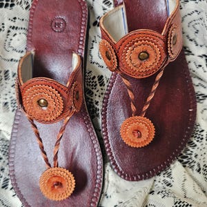 May include: A pair of brown leather sandals with decorative floral accents. The sandals feature a thong design with a circular, braided strap and a matching floral embellishment. The footbed is a darker shade of brown, and the sandals are set on a white lace surface.