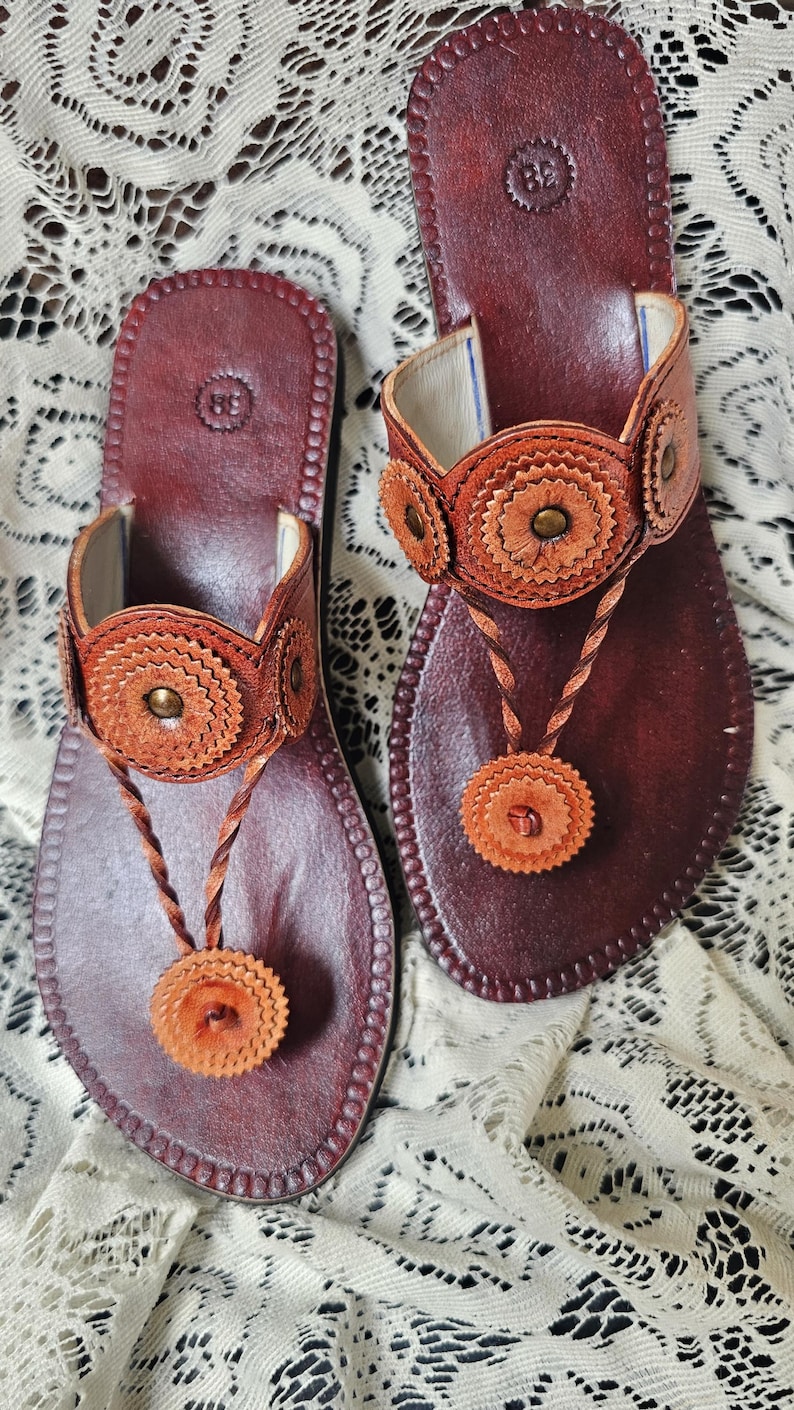 May include: A pair of brown leather sandals with decorative floral accents. The sandals feature a thong design with a circular, braided strap and a matching floral embellishment. The footbed is a darker shade of brown, and the sandals are set on a white lace surface.