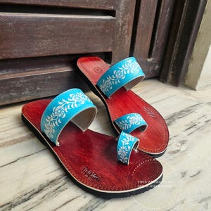 May include: A pair of leather sandals with a red footbed and black sole. The sandals feature light blue straps adorned with a white floral pattern. The sandals are handmade.