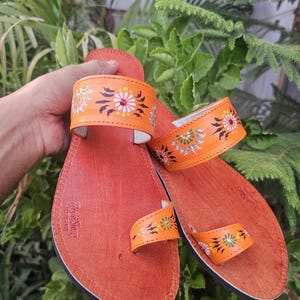 Op de afbeelding: Oranje leren sandalen met bloemmotieven in wit, groen en geel. De sandalen hebben een teengreep en een band over de bovenkant van de voet. De zolen zijn roodbruin.