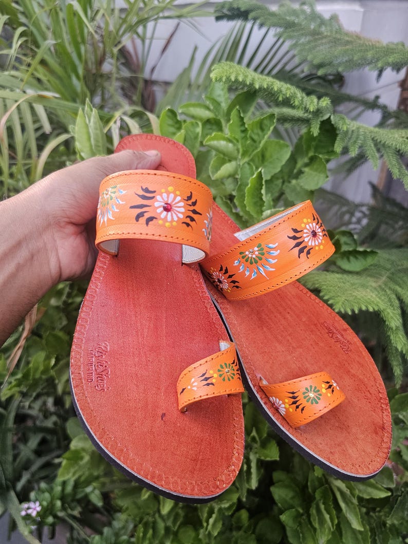 Op de afbeelding: Oranje leren sandalen met bloemmotieven in wit, groen en geel. De sandalen hebben een teengreep en een band over de bovenkant van de voet. De zolen zijn roodbruin.