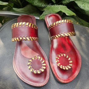 May include: Burgundy leather sandals with gold-coloured detailing. The sandals have a toe loop and a strap across the foot, both with gold accents. The brand name "leathereyes" is visible in the image.