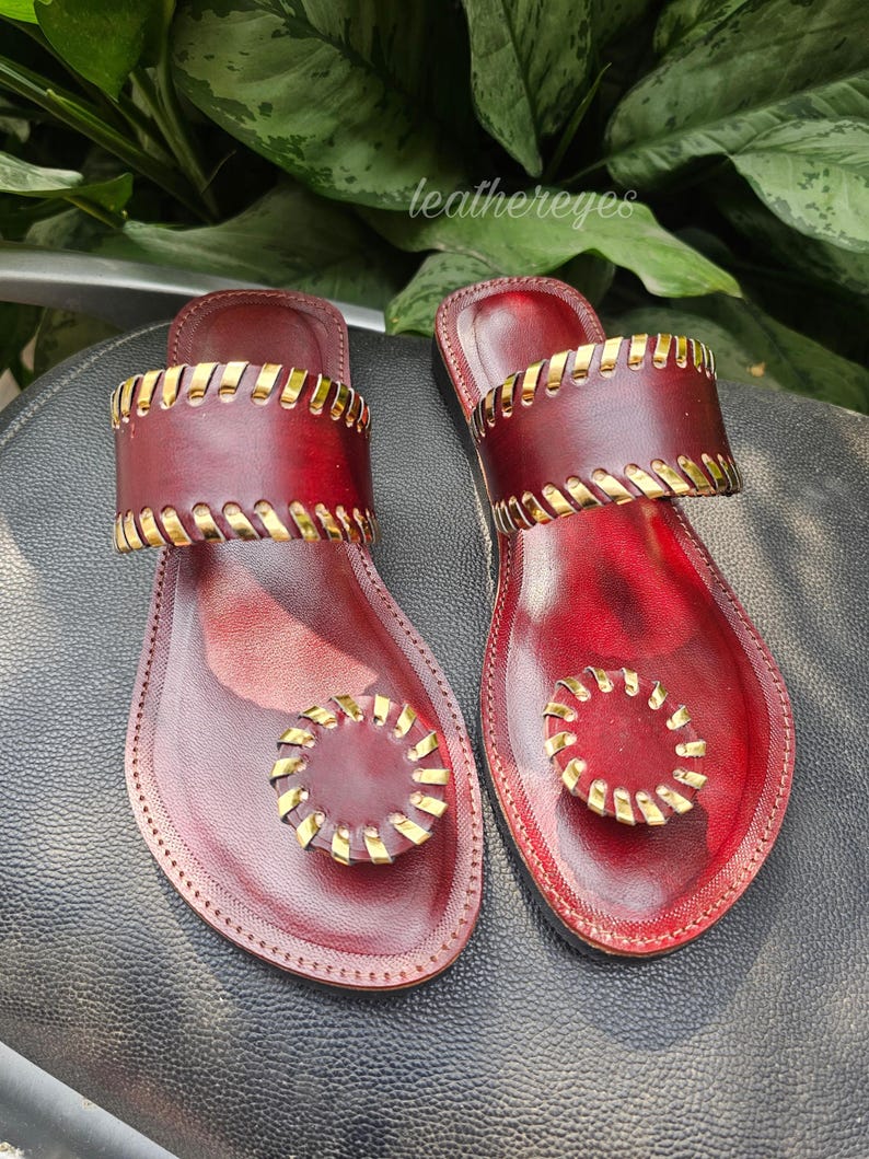 May include: Burgundy leather sandals with gold-coloured detailing. The sandals have a toe loop and a strap across the foot, both with gold accents. The brand name "leathereyes" is visible in the image.