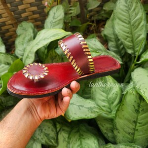 May include: Red leather sandals with gold detailing. The sandals have a circular embellishment on the toe strap and a gold-stitched strap. The sole is black. The sandals are displayed against a background of green leaves.