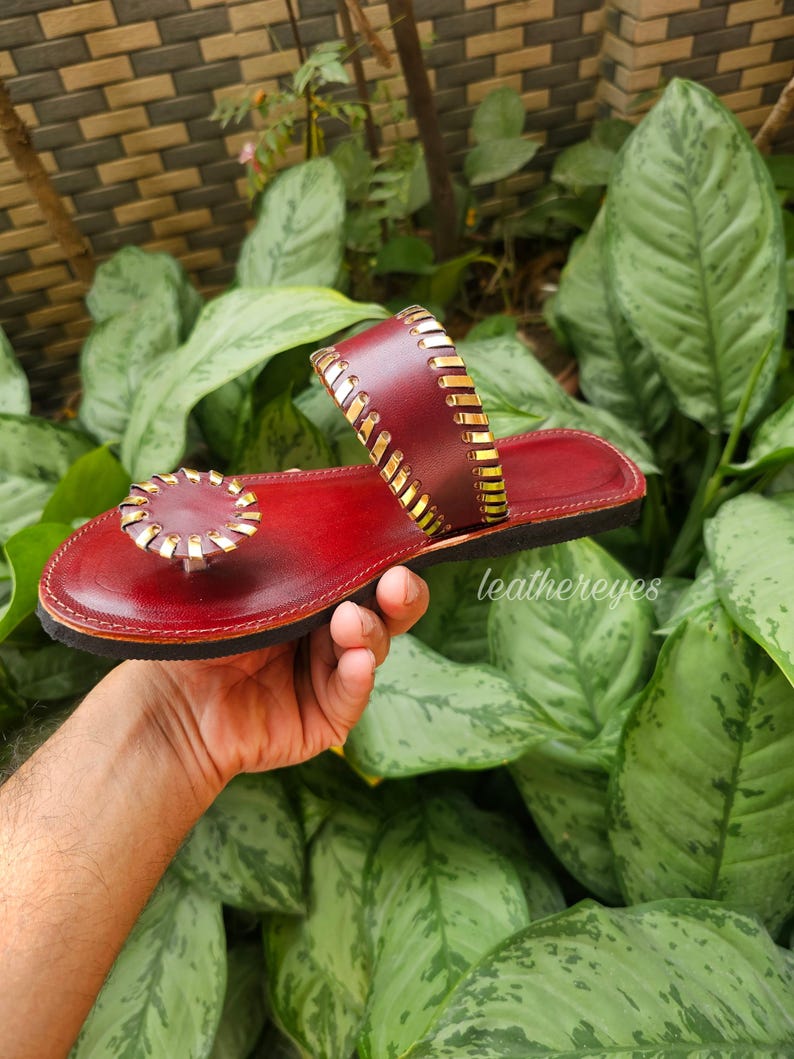 May include: Red leather sandals with gold detailing. The sandals have a circular embellishment on the toe strap and a gold-stitched strap. The sole is black. The sandals are displayed against a background of green leaves.
