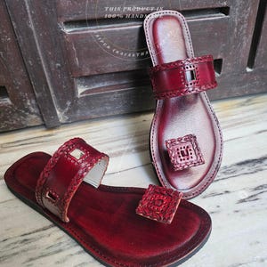 May include: A pair of handcrafted, burgundy leather sandals. The sandals feature a toe loop and ankle strap with decorative stitching and cut-out details. The soles are dark brown, and the sandals are displayed on a marble surface.