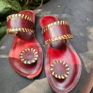 May include: Burgundy leather sandals with gold-coloured detailing. The sandals have a wide strap and a circular design on the toe strap. The sandals are displayed on a dark surface. The sandals are a stylish footwear option.