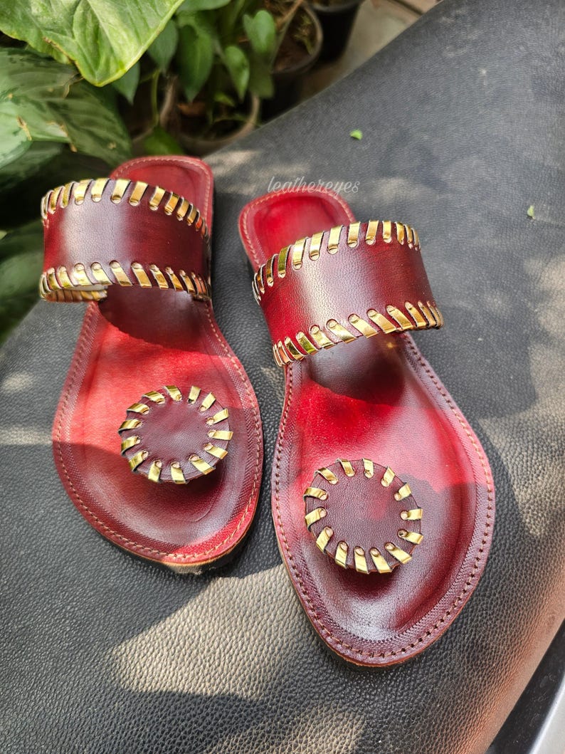 May include: Burgundy leather sandals with gold-coloured detailing. The sandals have a wide strap and a circular design on the toe strap. The sandals are displayed on a dark surface. The sandals are a stylish footwear option.
