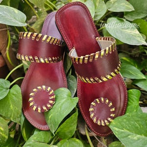 May include: Burgundy leather sandals with gold detailing. The sandals have a two-strap design with decorative stitching and a circular embellishment on the toe. The sandals are set against a backdrop of green leaves.