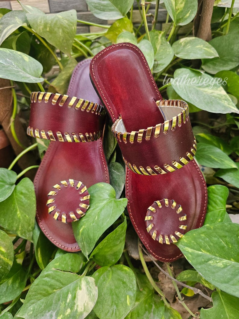 May include: Burgundy leather sandals with gold detailing. The sandals have a two-strap design with decorative stitching and a circular embellishment on the toe. The sandals are set against a backdrop of green leaves.
