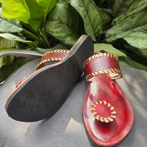 May include: Red leather sandals with gold stitching and accents. The sandals have a flat sole and a thong design. The brand name "leathereyes" is visible in the background. The sandals are displayed on a black surface.