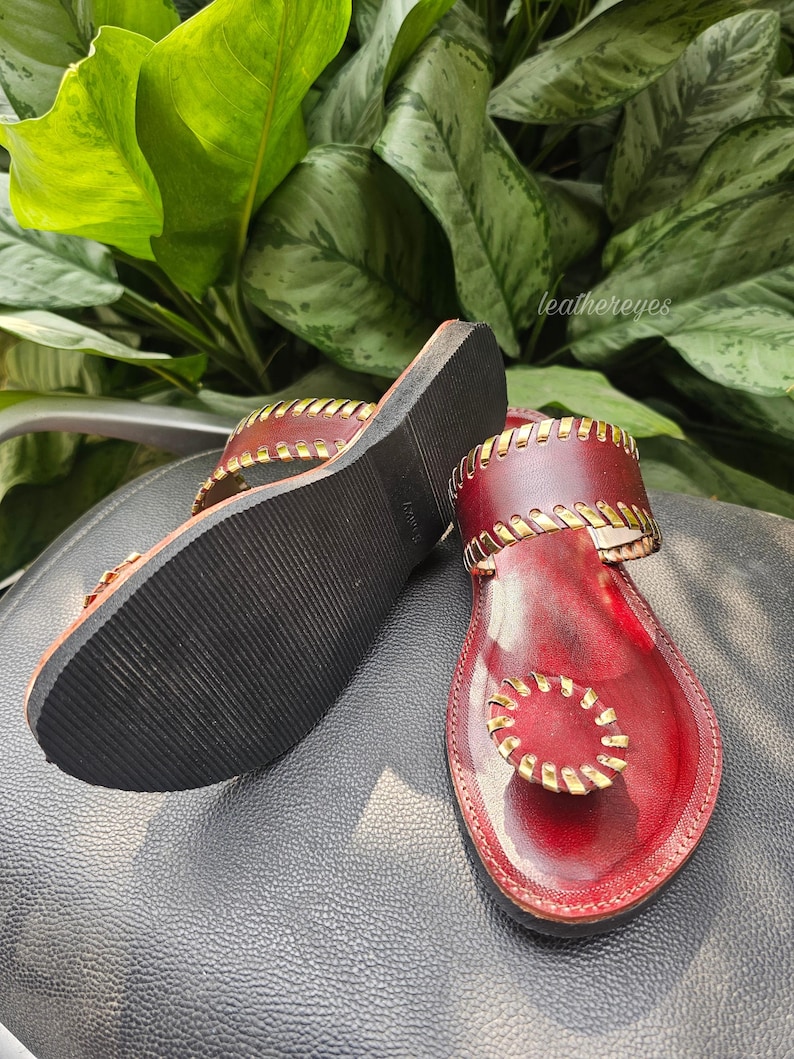 May include: Red leather sandals with gold stitching and accents. The sandals have a flat sole and a thong design. The brand name "leathereyes" is visible in the background. The sandals are displayed on a black surface.