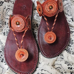 May include: A pair of handcrafted leather sandals. The sandals feature a deep burgundy footbed with intricate orange and tan floral accents. The thong strap and ankle strap are adorned with decorative elements and brass studs.