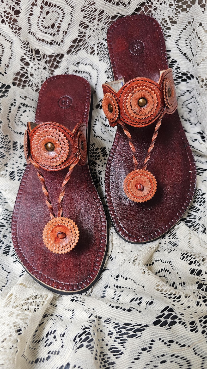 May include: A pair of handcrafted leather sandals. The sandals feature a deep burgundy footbed with intricate orange and tan floral accents. The thong strap and ankle strap are adorned with decorative elements and brass studs.