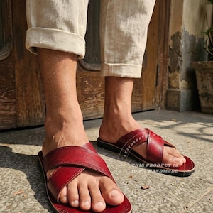 May include: Red leather sandals with a criss-cross strap design. The sandals have a dark sole and are handmade. The image shows the sandals being worn with beige pants.