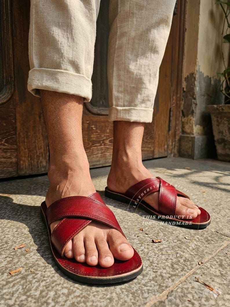 May include: Red leather sandals with a criss-cross strap design. The sandals have a dark sole and are handmade. The image shows the sandals being worn with beige pants.