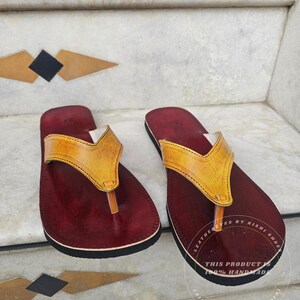 Pode incluir: Um par de sand&aacute;lias de couro artesanais. As sand&aacute;lias apresentam uma palmilha bord&ocirc; escura com sola preta e uma tira de couro amarela. O texto na sola diz "LEATHER EYES BY RISHI SHOE" e "THIS PRODUCT IS 100% HANDMADE JAIPUR (RAJASTHAN, INDIA)".
