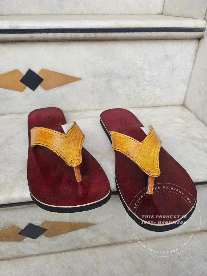 Pode incluir: Um par de sand&aacute;lias de couro artesanais. As sand&aacute;lias apresentam uma palmilha bord&ocirc; escura com sola preta e uma tira de couro amarela. O texto na sola diz "LEATHER EYES BY RISHI SHOE" e "THIS PRODUCT IS 100% HANDMADE JAIPUR (RAJASTHAN, INDIA)".