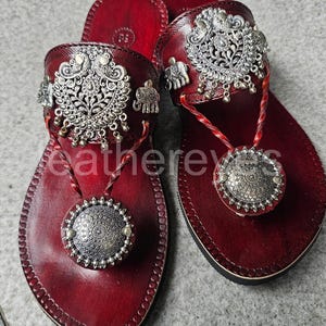 Könnte beinhalten: Ein Paar burgunderfarbene Ledersandalen mit silbernen Verzierungen. Die Sandalen haben aufwendige silberne Designs auf den Riemen und Zehenstegen, mit kleinen Elefanten-Anhängern. Ein großes silbernes Medaillon ist am Zehensteg befestigt.