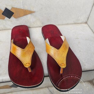 Pode incluir: Um par de sand&aacute;lias de couro feitas &agrave; m&atilde;o. As sand&aacute;lias t&ecirc;m uma palmilha vermelha escura com uma tira de couro amarela. A tira tem formato de V. As sand&aacute;lias s&atilde;o rotuladas como "LEATHEREYES BY RISH SHOES" e "THIS PRODUCT IS 100% HANDMADE JAIPUR (RAJASTHAN, INDIA)".