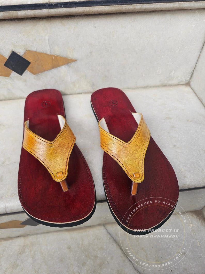 Pode incluir: Um par de sand&aacute;lias de couro feitas &agrave; m&atilde;o. As sand&aacute;lias t&ecirc;m uma palmilha vermelha escura com uma tira de couro amarela. A tira tem formato de V. As sand&aacute;lias s&atilde;o rotuladas como "LEATHEREYES BY RISH SHOES" e "THIS PRODUCT IS 100% HANDMADE JAIPUR (RAJASTHAN, INDIA)".