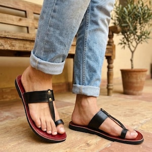 Peut inclure: Une paire de sandales entre-doigts en cuir noir avec une boucle d'orteil, une large bride horizontale fixée par deux petits rivets métalliques dorés et une bordure de semelle intérieure rouge foncé, portées avec un jean bleu clair à revers.