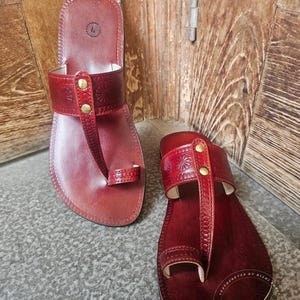 May include: A pair of handmade, red leather sandals. The sandals feature a toe loop and a strap across the top of the foot, adorned with decorative stitching and brass-colored studs. The soles are dark brown.