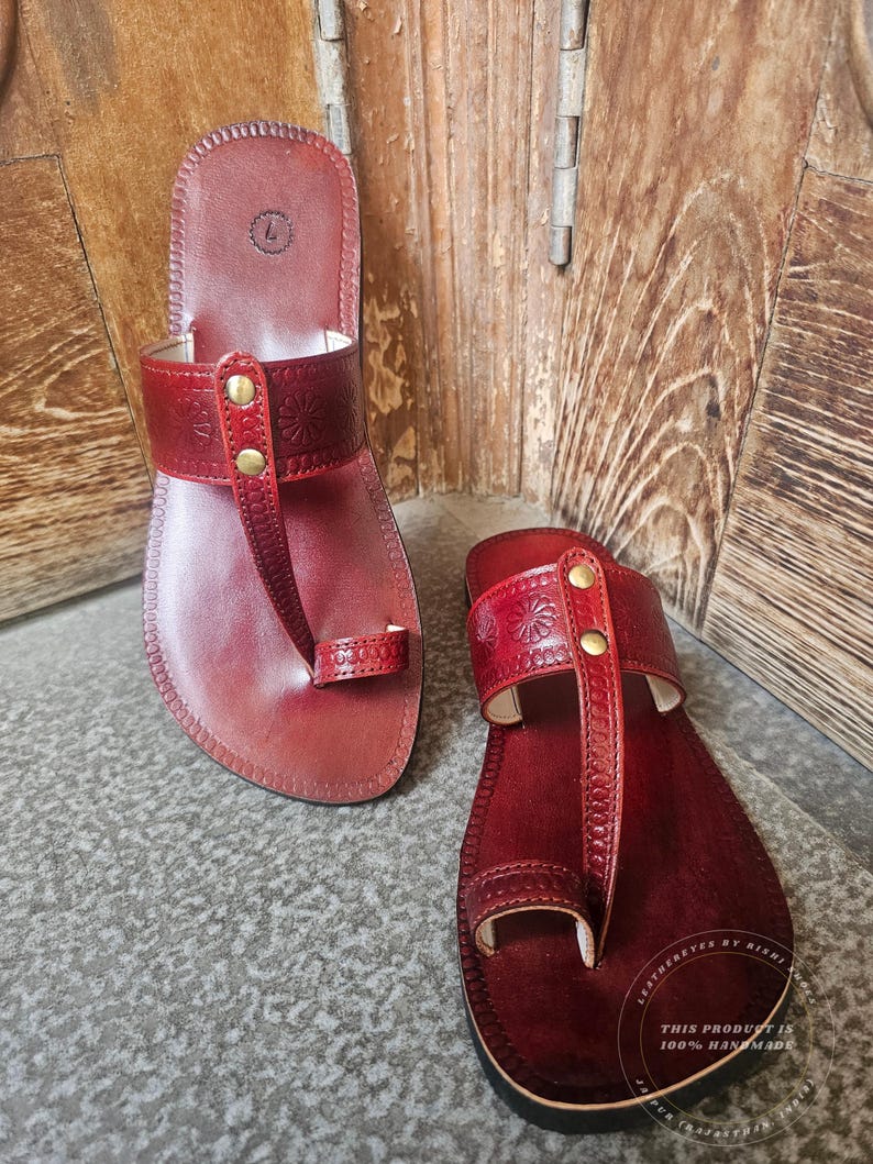 May include: A pair of handmade, red leather sandals. The sandals feature a toe loop and a strap across the top of the foot, adorned with decorative stitching and brass-colored studs. The soles are dark brown.